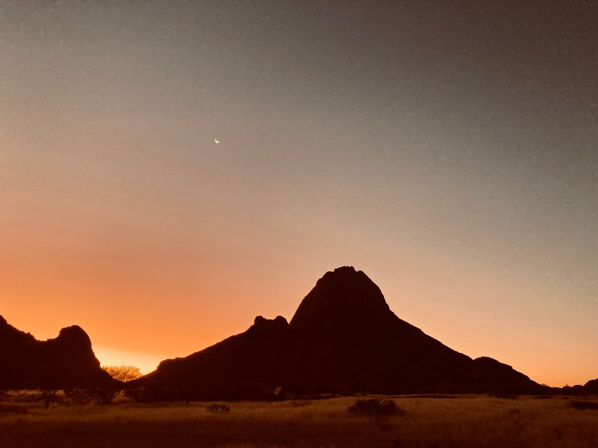 Spitzkoppe silhouetted against an orange sunset sky with a crescent moon