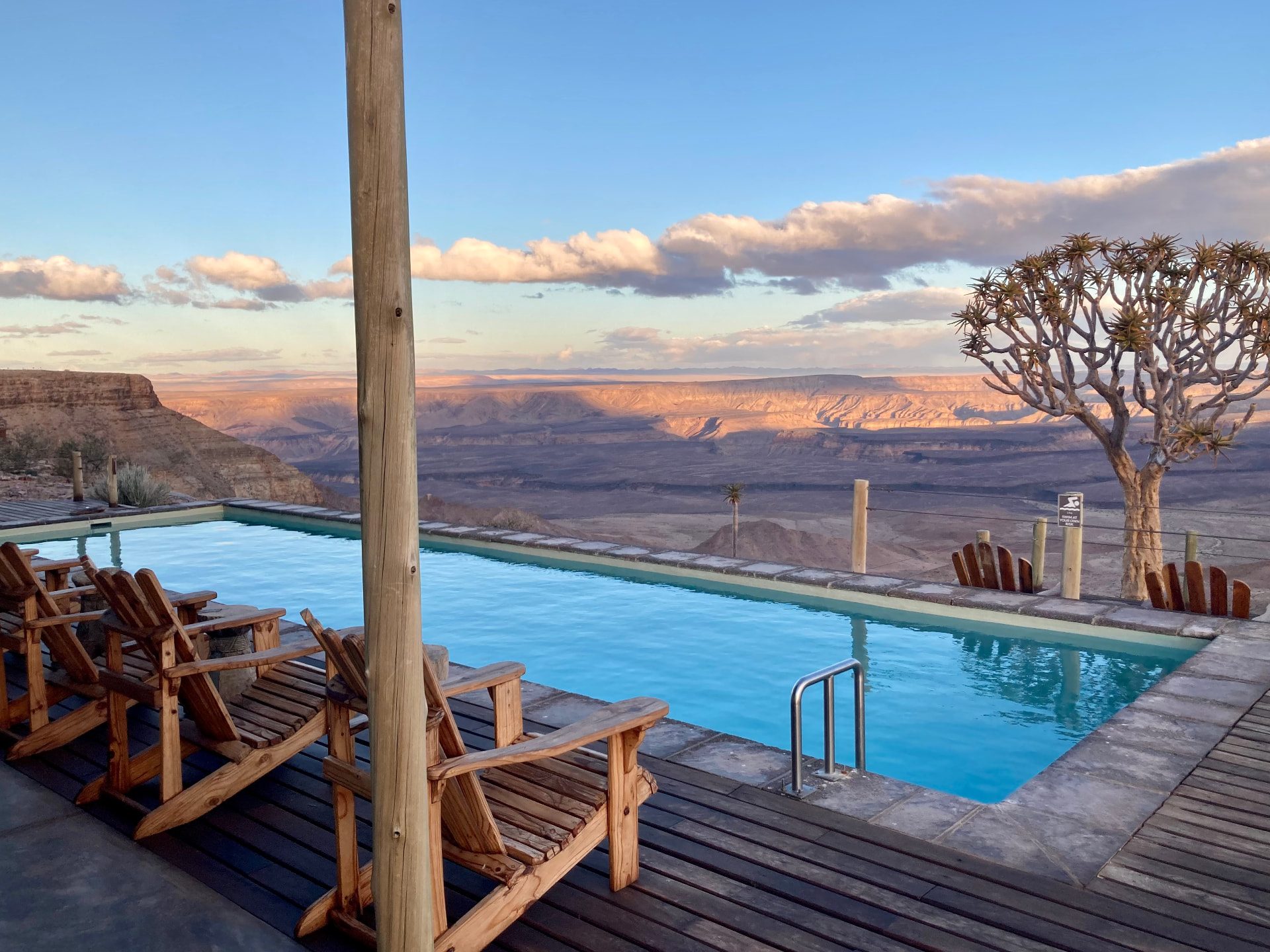An infinity pool and wooden loungers on a lodge deck overlooking Fish River Canyon at sunset, a quiver tree alongside