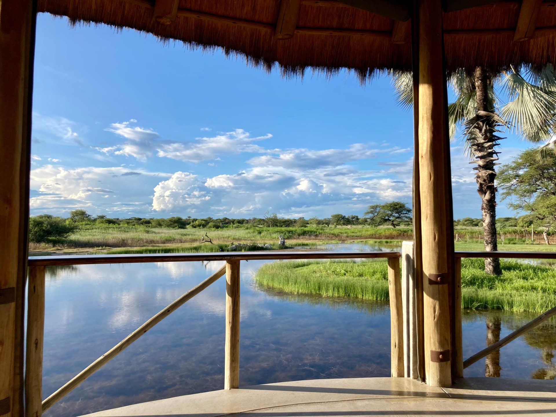 View from a thatched lodge deck across a still waterhole to green grassland and a big cloud-filled sky