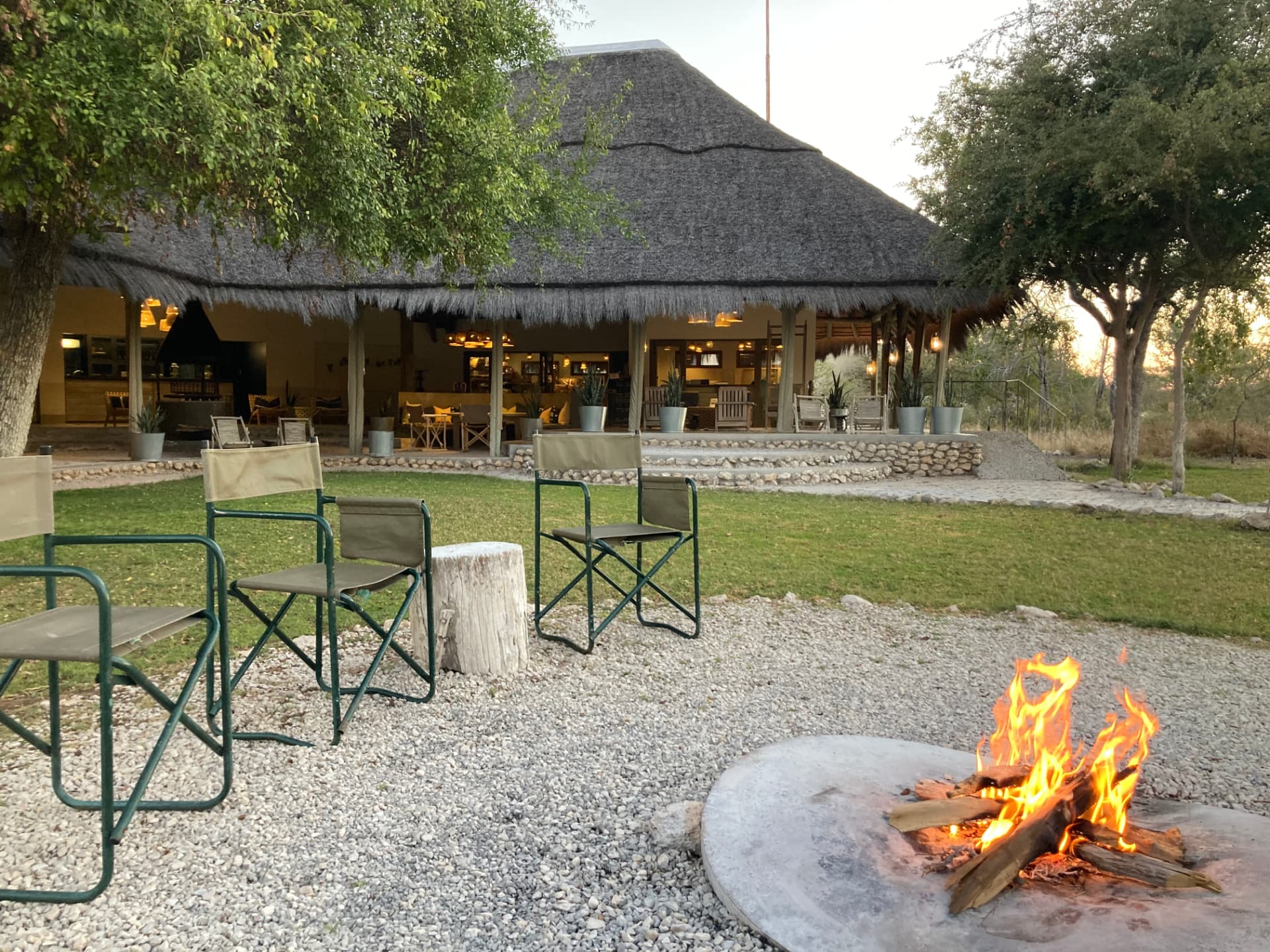 Safari chairs around a lit fire pit on gravel in front of a thatched lodge at dusk